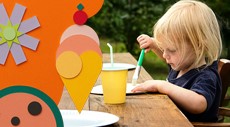 Preschool child painting at outdoor craft table with colorful paper summer decorations including ice cream, flowers, and shapes